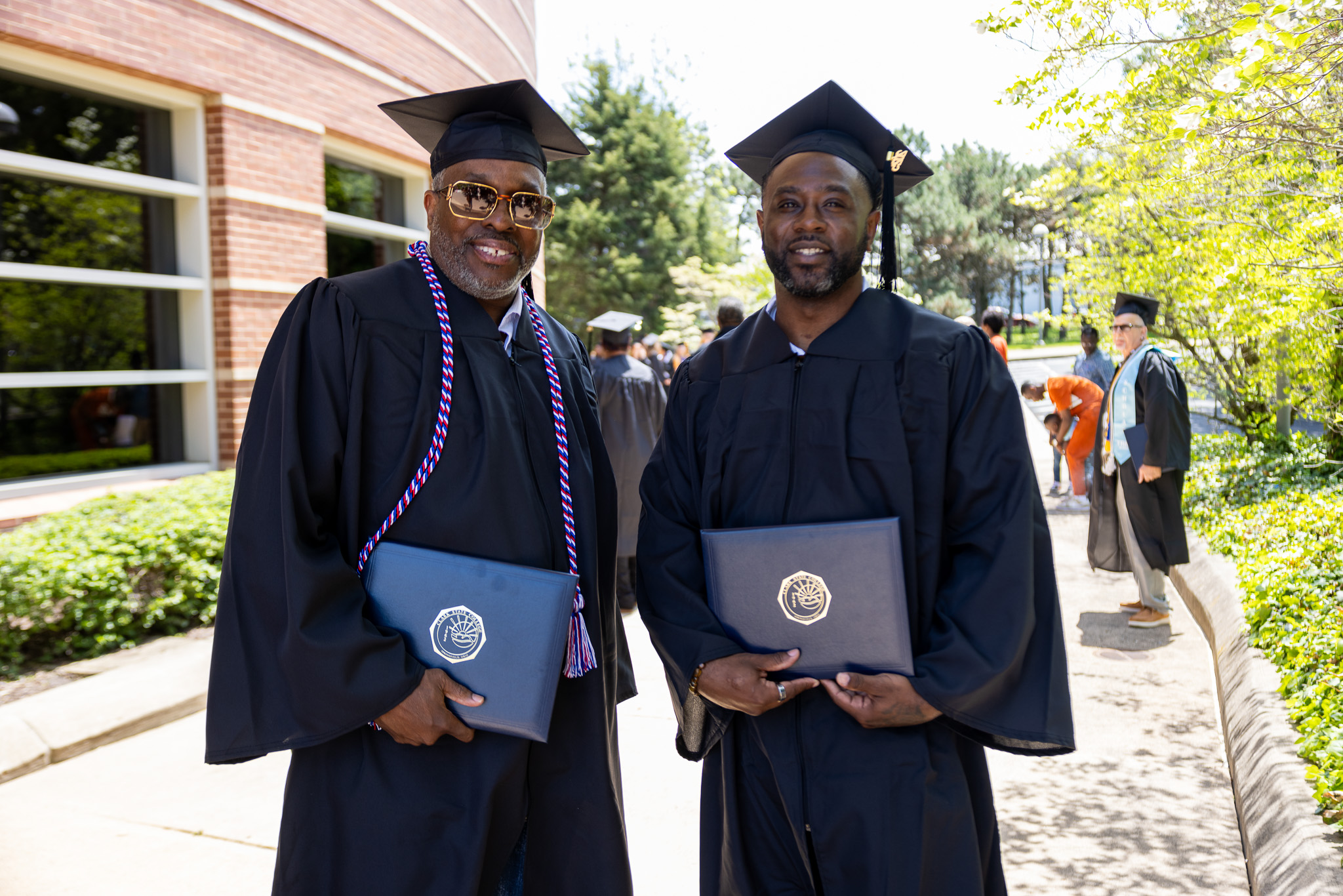 Two graduates in black caps and gowns smile outdoors, each holding a diploma folder, with a campus building and other graduates in the background.